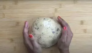 Hands holding wrapped chocolate chip cookie dough on a wooden surface, ready for baking.